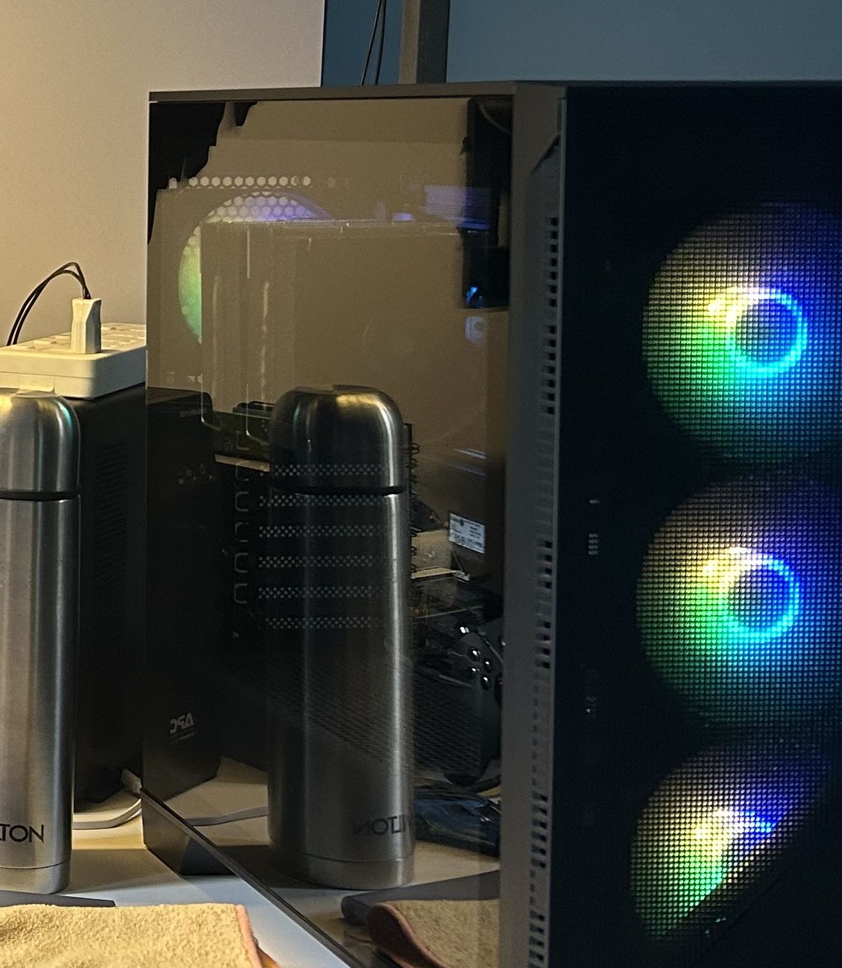 A glass-panelled computer tower glowing with RGB-lit fans, part of a home server setup, beside a UPS battery backup — the corner of a room running like a small data centre