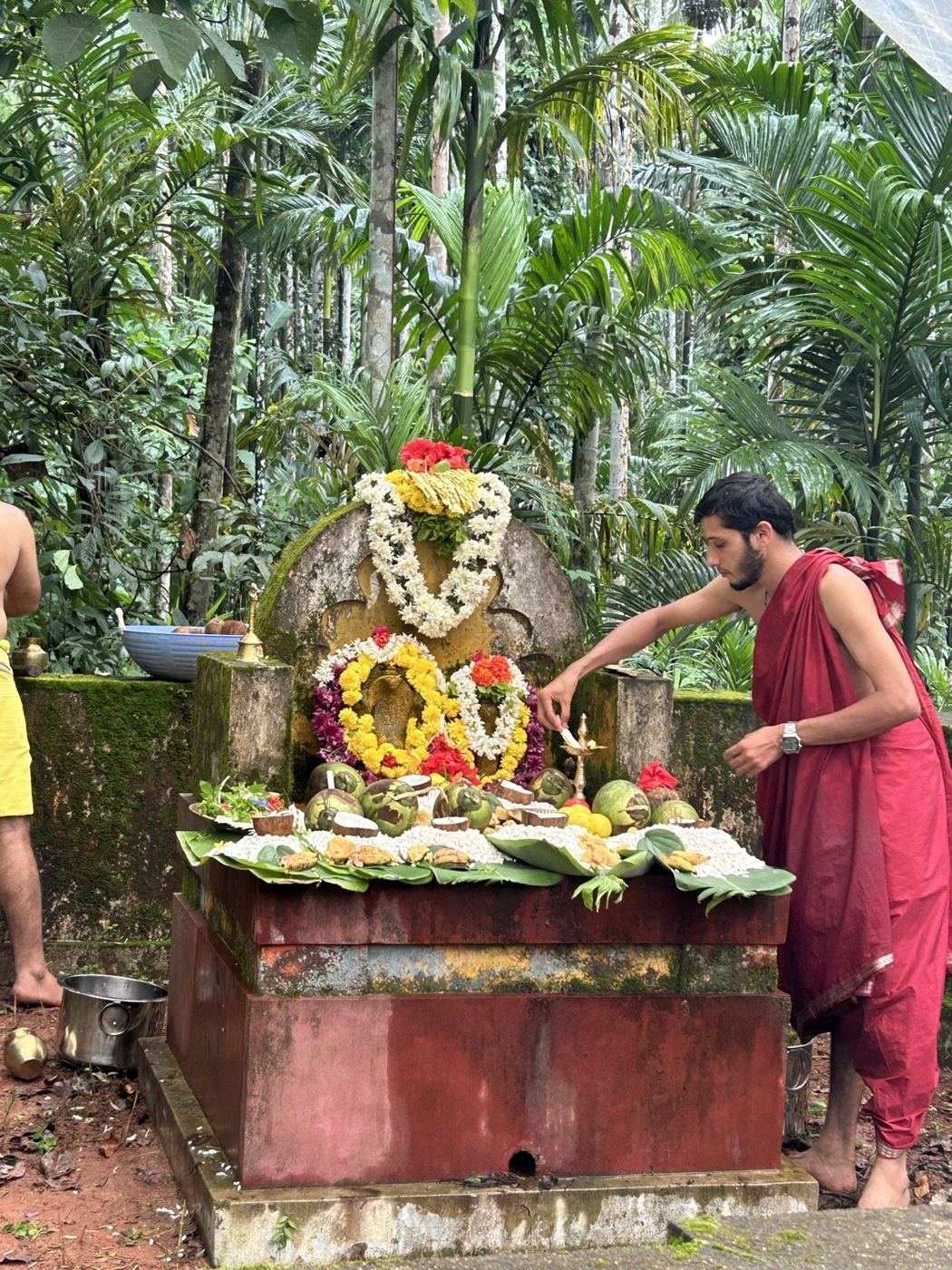 A garlanded naga stone — a carved serpent deity — decorated with marigold and jasmine garlands, with offerings of coconuts, fruit and puffed rice on banana leaves, during Nagara Panchami puja in a forest grove